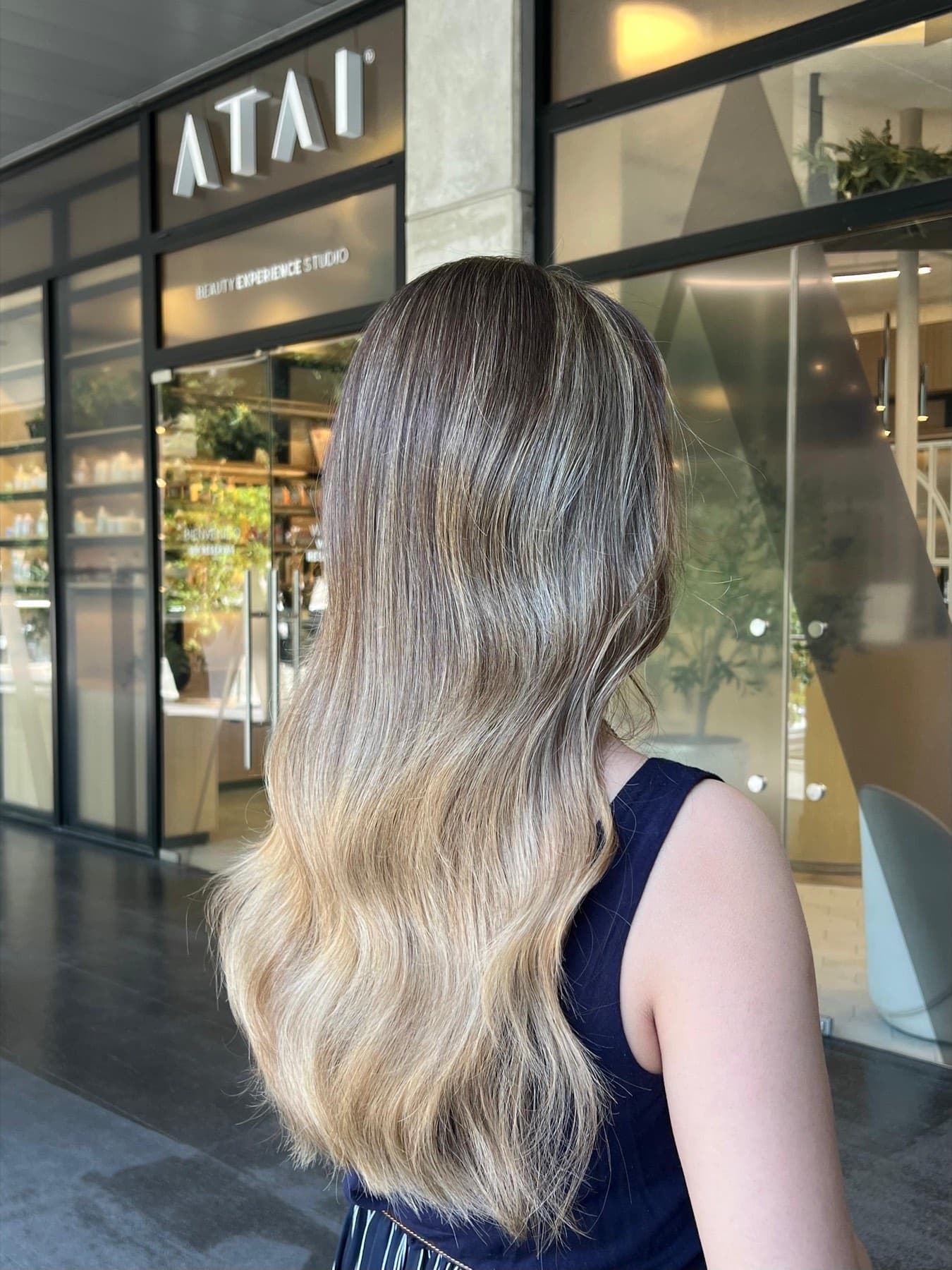 Melena larga con balayage rubio, de espalda, frente a la fachada del salón Atai